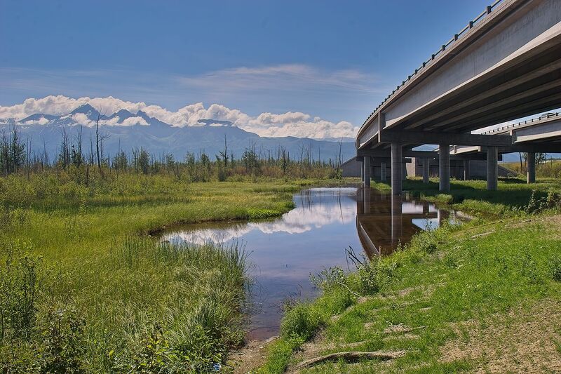 Still water winding through the Palmer Hay Flats beneath the Glenn Highway overpass with the Chugach mountains behind